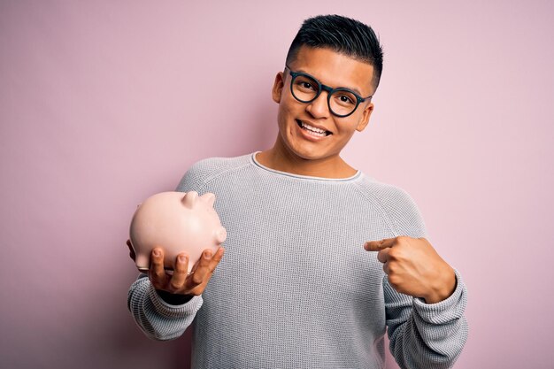 Junger gutaussehender lateinamerikanischer Mann mit Sparschwein über isoliertem rosa Hintergrund mit überraschendem Gesicht, das mit dem Finger auf sich selbst zeigt