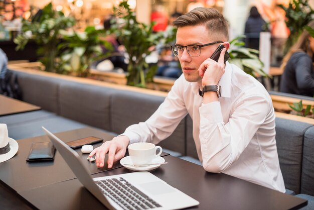 Junger gutaussehender Mann, der am Handy im Café mit Laptop auf Tabelle spricht