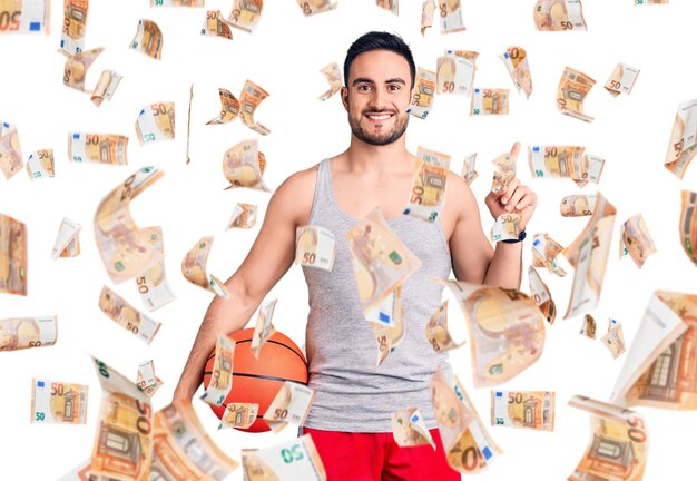Junger gutaussehender mann, der basketballball hält, überrascht mit einer idee oder frage, die finger mit glücklichem gesicht nummer eins zeigt
