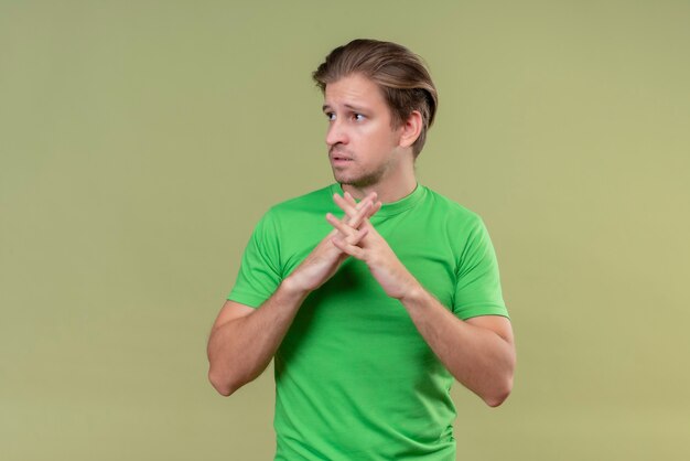 Junger gutaussehender Mann, der grünes T-Shirt hält, das Hände zusammenhält und mit traurigem Ausdruck auf Gesicht steht, das über grüner Wand steht