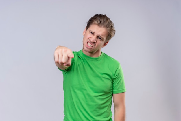 Junger gutaussehender Mann, der grünes T-Shirt trägt, das mit dem Finger zur Kamera zeigt, die mit verärgertem Ausdruck auf Gesicht steht, das über weißer Wand steht unzufrieden aussieht