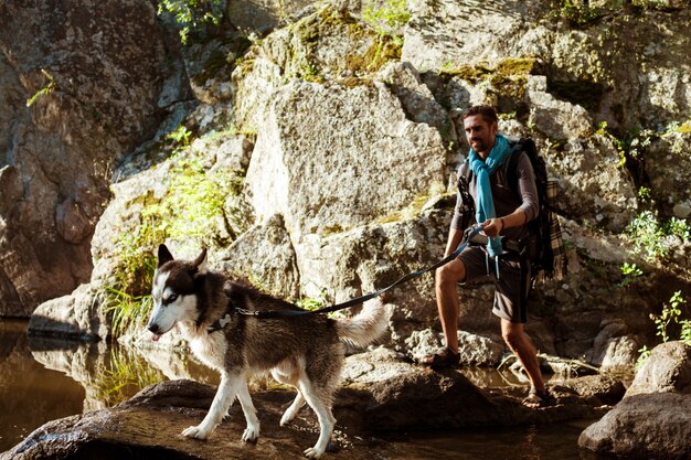 Junger gutaussehender Mann, der mit Huskys Hund in der Schlucht nahe Wasser geht