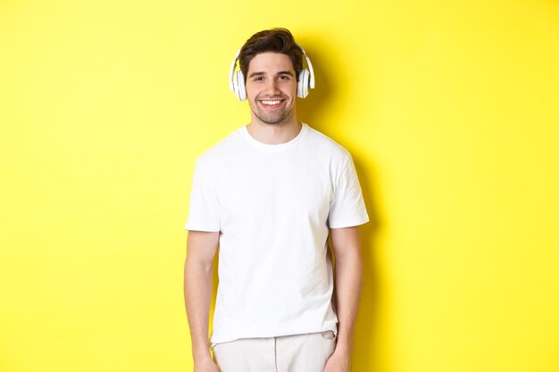 Junger gutaussehender Mann, der Musik in Kopfhörern hört, Kopfhörer trägt und lächelt, über gelbem Hintergrund stehend.