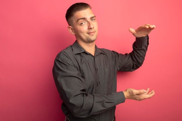 Kostenloses Foto junger gutaussehender mann im grauen hemd, das vorne lächelnd selbstbewusst zeigt größe geste mit händen maß symbol steht über rosa wand