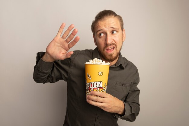 Junger gutaussehender Mann im grauen Hemd, der Eimer mit Popcorn hält, das besorgt und verwirrt beiseite schaut, die Hand heraus wie Verteidigungsgeste aushält