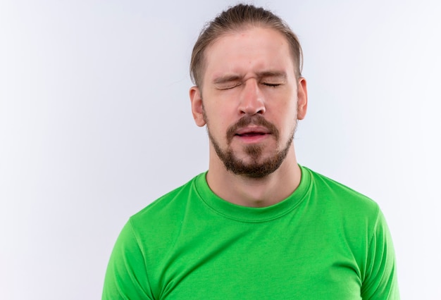 Junger gutaussehender Mann in den grünen T-Shirt-schließenden Augen erschrocken stehend über weißem Hintergrund