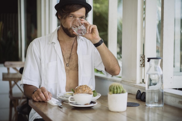 Junger gutaussehender Mann in einem weißen Hemd offen, Frühstück in einem Café mit vegetarischem Burger, Kaffeetrinken, Lebensstil auf einer tropischen Insel, Leben auf Bali.