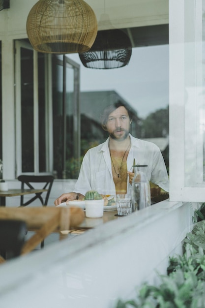 Junger gutaussehender Mann in einem weißen Hemd offen, Frühstück in einem Café mit vegetarischem Burger, Kaffeetrinken, Lebensstil auf einer tropischen Insel, Leben auf Bali.