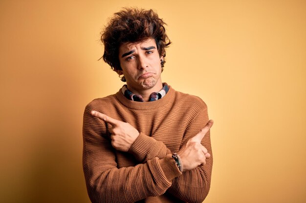 Junger, gutaussehender Mann in lässigem Hemd und Pullover vor isoliertem gelbem Hintergrund, der mit den Fingern in unterschiedliche Richtungen auf beide Seiten zeigt, stimmt nicht überein