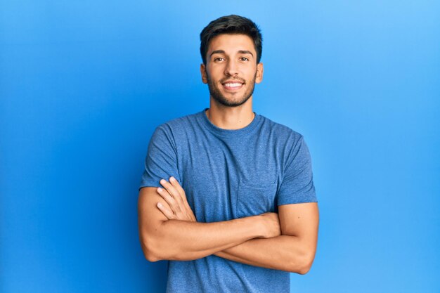 Junger gutaussehender Mann in lässigem T-Shirt vor blauem Hintergrund, glückliches Gesicht, lächelnd und mit verschränkten Armen in die Kamera blickend. positive Person.