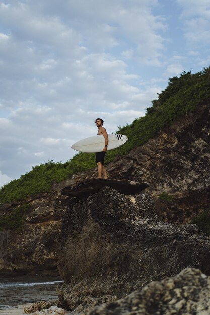 Junger gutaussehender Mann mit einem Surfbrett auf einem Felsen in der Nähe des Ozeans.