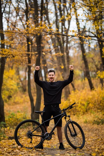 Junger gutaussehender Mannbiker nach dem Training mit erhobenen Händen im Herbstwaldweg