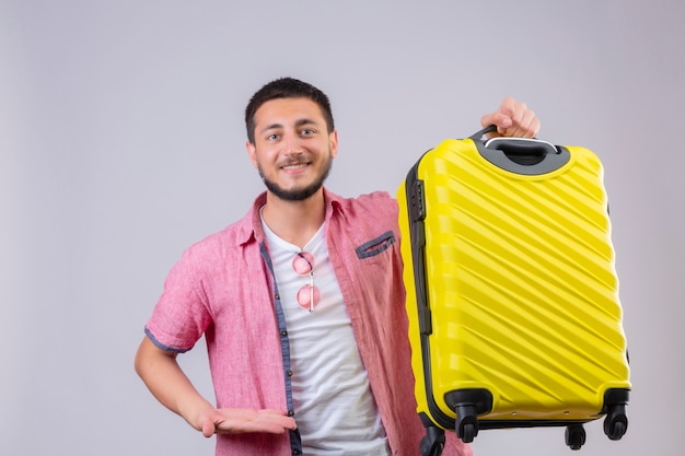 Junger gutaussehender Reisender Kerl, der Koffer hält Kamera mit zuversichtlichem Lächeln positiv und glücklich steht über weißem Hintergrund
