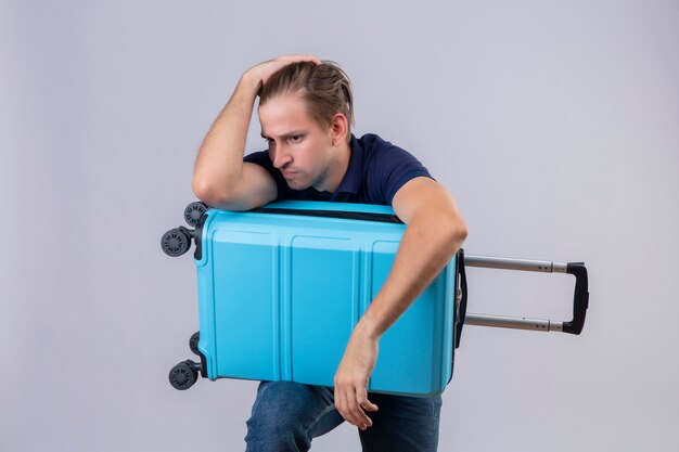 Junger gutaussehender Reisender Kerl, der mit Koffer steht, der stressig Hände auf Kopf müde und frustriert über weißem Hintergrund hält