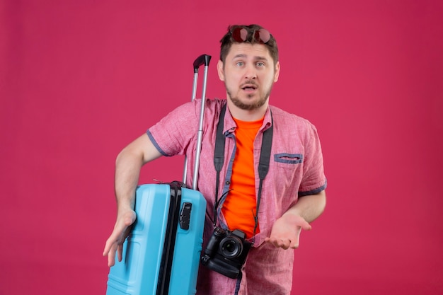 Junger gutaussehender reisender Mann mit der Kamera, die mit Koffer steht, der verwirrte achselzuckende Schultern schaut, die unsicher und verwirrt aussehen