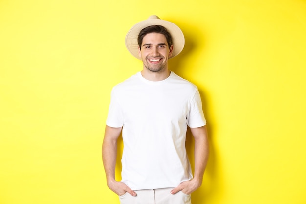 Junger gutaussehender Tourist, der glücklich aussieht, Strohhut zum Reisen trägt und vor gelbem Hintergrund steht