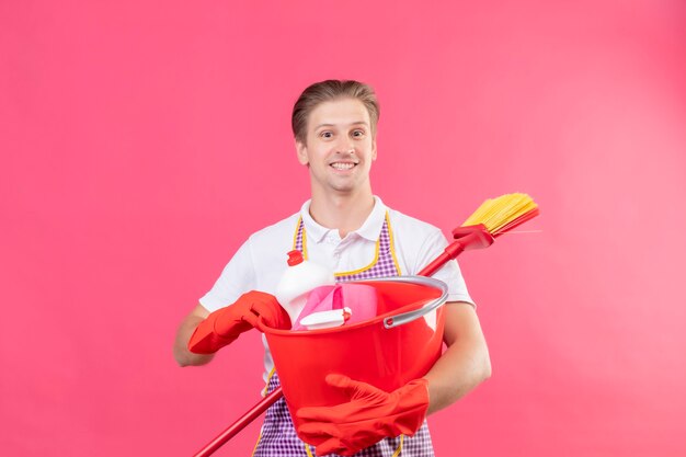 Junger hansdome Mann, der Schürze hält Eimer mit Reinigungswerkzeugen und Mopp lächelnd mit glücklichem Gesicht steht über rosa Wand