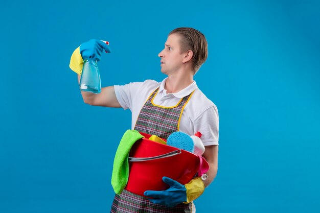 Junger Hansdome-Mann, der Schürze und Gummihandschuhe hält, die Eimer mit Reinigungswerkzeugen halten