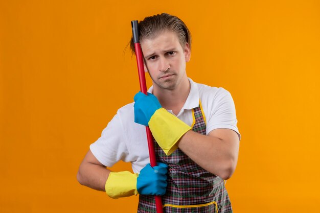 Junger Hansdome-Mann, der Schürze und Gummihandschuhe hält, die Mopp halten, der müde und überarbeitet mit traurigem Ausdruck sieht, der über orange Wand steht