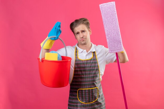 Junger Hansdome-Mann, der Schürze und Gummihandschuhe hält Eimer mit Reinigungswerkzeugen und Mopp hält