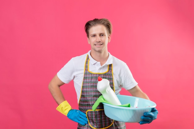 Junger Hansdome-Mann, der Schürze und Gummihandschuhe trägt, die Becken mit Reinigungswerkzeugen mit dem selbstbewussten Lächeln auf Gesicht halten, das über rosa Wand steht