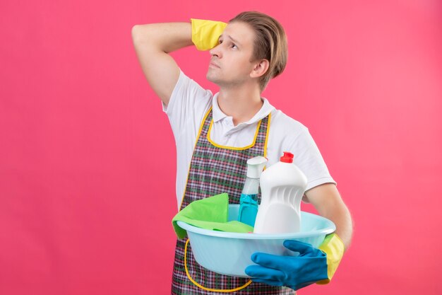 Junger Hansdome-Mann, der Schürze und Gummihandschuhe trägt, die Eimer mit Reinigungswerkzeugen halten, die mit nachdenklichem Ausdruck auf Gesicht denkend stehen und über rosa Wand stehen