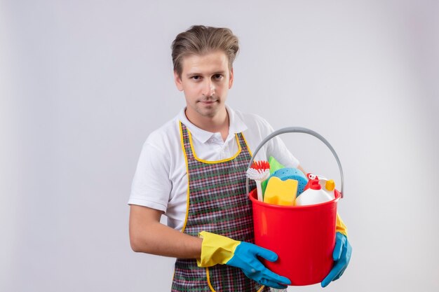 Junger Hansdome-Mann, der Schürze und Gummihandschuhe trägt, die Eimer mit Reinigungswerkzeugen mit sicherem Lächeln auf Gesicht halten, das über weißer Wand 2 steht