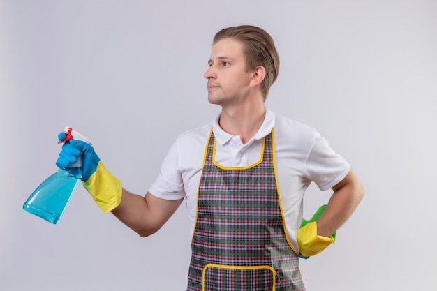 Junger Hansdome-Mann, der Schürze und Gummihandschuhe trägt, die Reinigungsspray und Teppich halten