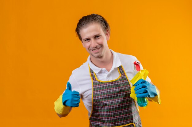 Junger Hansdome-Mann, der Schürze und Gummihandschuhe trägt, die Reinigungsspray und Teppich halten