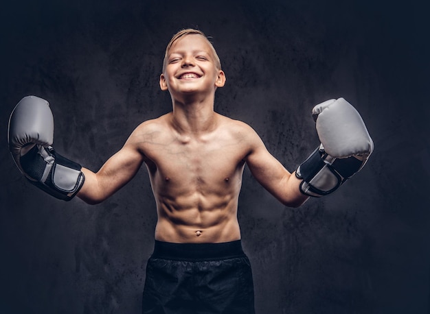 Junger hemdloser Boxer mit Boxhandschuhen hat Spaß beim Posieren in einem Studio. Getrennt auf einem dunklen strukturierten Hintergrund.