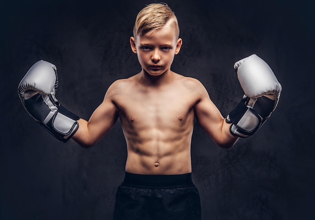 Junger hemdloser Boxer mit Boxhandschuhen posiert in einem Studio. Getrennt auf einem dunklen strukturierten Hintergrund.