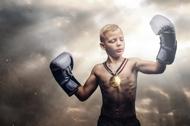 Junger hemdloser Champion-Junge mit Boxhandschuhen posiert auf dem Hintergrund des dunklen bewölkten Himmels, der die Sonnenstrahlen durchdringt.