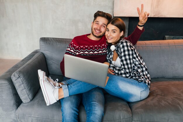 Junger Hipster lächelnder Mann und Frau, die zu Hause im Winter sitzen, Laptop halten, Paar auf Freizeit zusammen, online studierend, freiberuflich, glücklich, datierend, Daumen hoch zeigend, positiv, in der Kamera schauend