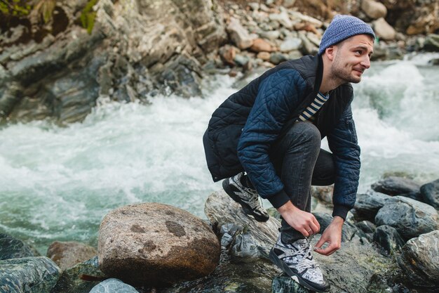 Junger Hipster-Mann, der auf einem Felsen am Fluss im Winterwald geht und Schnürsenkel bindet