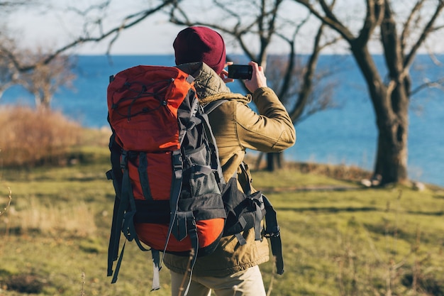 Junger Hipster-Mann, der mit dem Rucksack reist, der warme Jacke und Hut trägt, aktiver Tourist, Fotos auf Handy macht, Natur in der kalten Jahreszeit erforschend