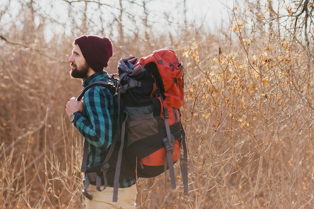 Junger Hipster-Mann, der mit Rucksack im Herbstwald mit kariertem Hemd und Hut reist, aktiver Tourist, Natur in der kalten Jahreszeit erforschend