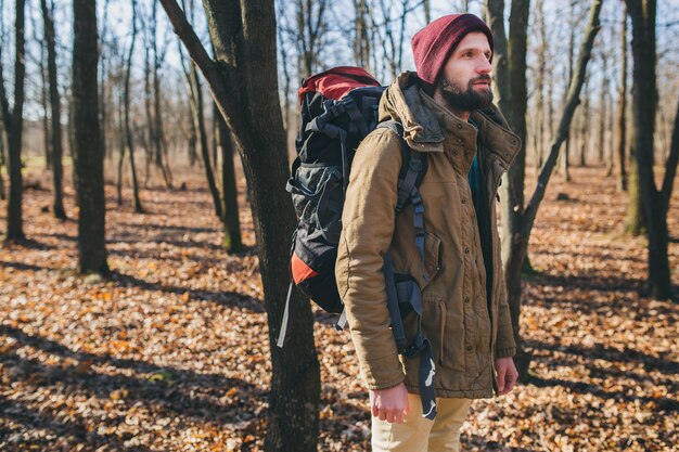 Junger Hipster-Mann, der mit Rucksack im Herbstwald mit warmer Jacke und Hut reist, aktiver Tourist, Natur in der kalten Jahreszeit erforschend