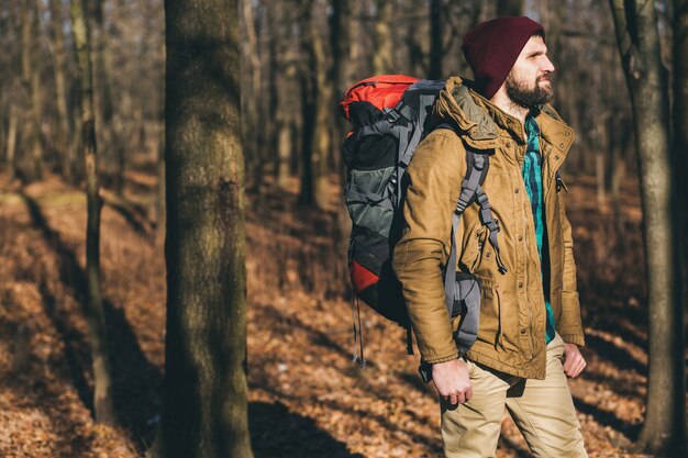 Junger Hipster-Mann, der mit Rucksack im Herbstwald mit warmer Jacke und Hut reist, aktiver Tourist, Natur in der kalten Jahreszeit erforschend