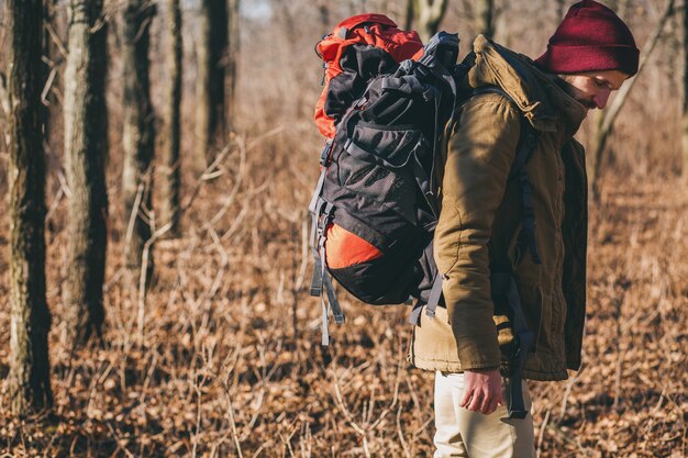 Junger Hipster-Mann, der mit Rucksack im Herbstwald mit warmer Jacke und Hut reist, aktiver Tourist, Natur in der kalten Jahreszeit erforschend