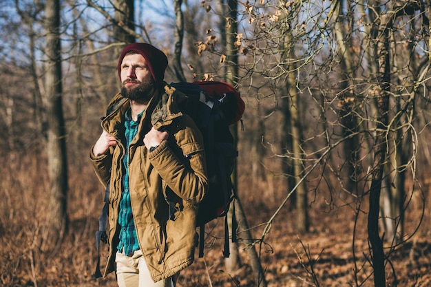 Junger Hipster-Mann, der mit Rucksack im Herbstwald mit warmer Jacke und Hut reist, aktiver Tourist, Natur in der kalten Jahreszeit erforschend