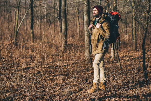 Junger Hipster-Mann, der mit Rucksack im Herbstwald mit warmer Jacke und Hut reist, aktiver Tourist, Natur in der kalten Jahreszeit erforschend