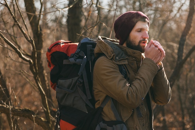 Junger Hipster-Mann, der mit Rucksack im Herbstwald reist und warme Jacke und Hut trägt