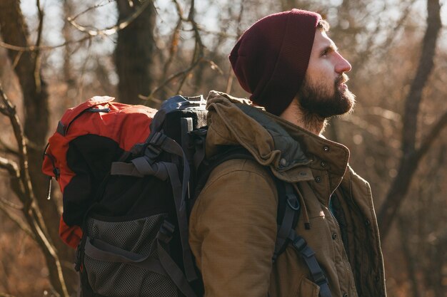 Junger Hipster-Mann, der mit Rucksack im Herbstwald reist und warme Jacke und Hut trägt