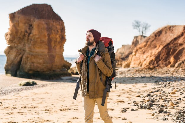 Junger Hipster-Mann, der mit Rucksack in der Herbstküste reist und warme Jacke und Hut trägt