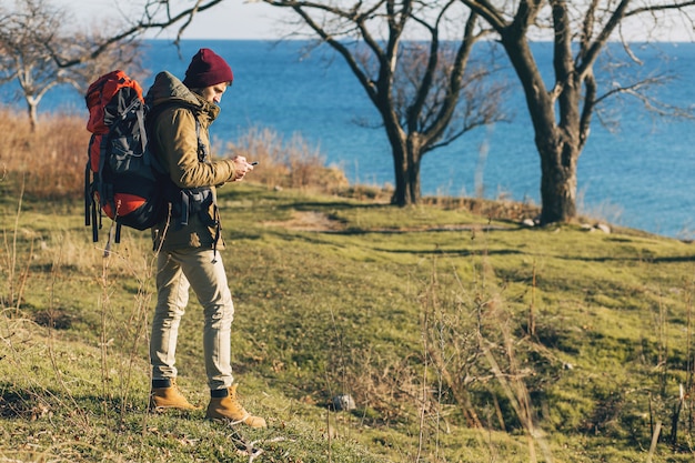 Junger Hipster-Mann, der mit Rucksack reist, der warme Jacke und Hut trägt, aktiver Tourist, mit Handy, Erkundung der Natur in der kalten Jahreszeit