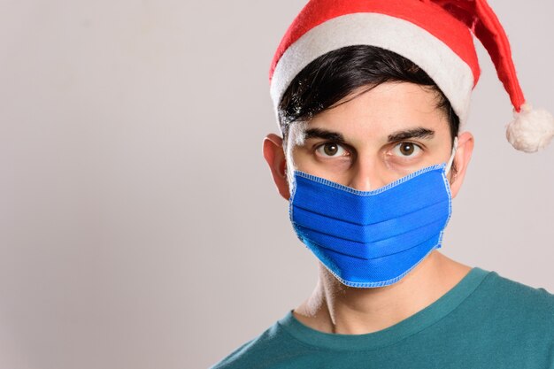 Junger hispanischer Mann, der eine Gesichtsmaske und eine Weihnachtsmannmütze auf weißem Hintergrund trägt