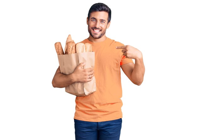 Junger hispanischer Mann, der eine Papiertüte mit Brot hält und mit dem Finger auf ein Selbst zeigt, das glücklich und stolz lächelt