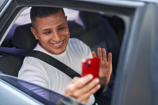 Junger hispanischer Mann, der im Auto einen Videoanruf mit dem Smartphone macht, sieht positiv und glücklich aus, steht und lächelt mit einem selbstbewussten Lächeln, das Zähne zeigt