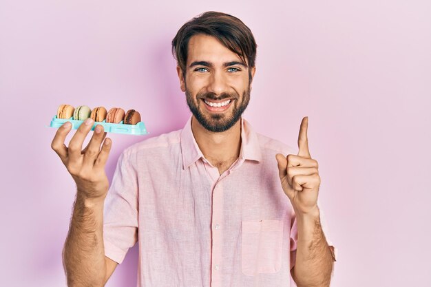 Junger hispanischer Mann, der köstliches Makrongebäck hält und mit einer Idee oder Frage lächelt und mit dem Finger mit glücklichem Gesicht zeigt, Nummer eins