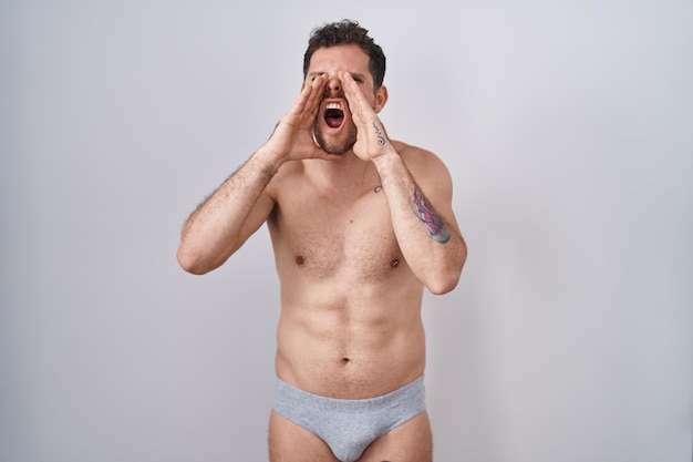 Junger hispanischer Mann, der mit nacktem Oberkörper steht und Unterwäsche trägt und wütend laut mit den Händen über dem Mund schreit
